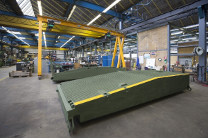 Ein große Lagerhalle mit zahlreichen grünen Behältern auf dem Boden, verschiedenen Maschinen und einer Wand im Hintergrund, die eine Uhr und angeklebte Papiere zeigt.