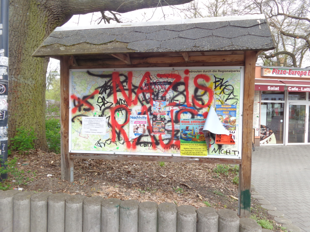 Ein Obdach mit Informationsschildern, Plakate an einem Pfahl, ein Gebäude mit Glastüren, Bäume, verstreute Blätter und ein Graffiti-Schild davor.