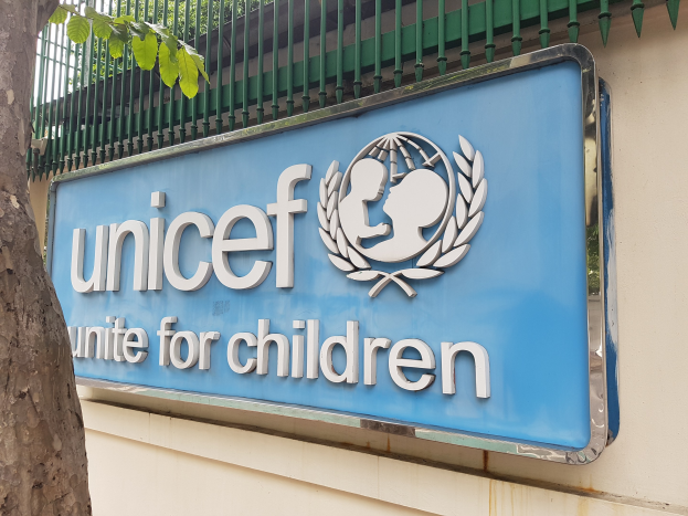 UNICEF-Logo auf einer Gebäudewand mit einem Baumstamm links und einer Tafel an der Wand, Bäume im Hintergrund unter einem klaren blauen Himmel.