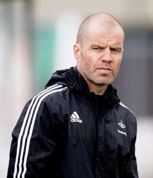 Baldiger Trainer in marineblauem Jackett mit weißen Streifen und weißem Kragen auf einem Fußballfeld stehend, Hände verschränkt, ernster Gesichtsausdruck, unscharfer Hintergrund.