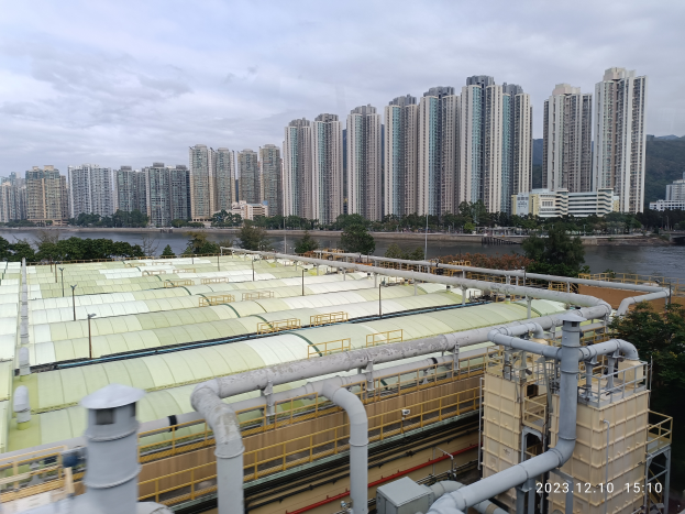Eine Stadtpanoramaview von einem Gebäude aus, mit Gebäuden, Bäumen, Pfählen, Wasser im Vordergrund und einem bewölkten Himmel im Hintergrund, mit einem Wasserzeichen, das eine Kläranlage in Hong Kong anzeigt.