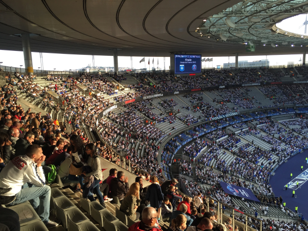 Große Zuschauermenge in einem Stadion bei einem Fußballspiel, mit einer Bühne rechts, Fahnen, Stangen, einem Bildschirm und der Allianz Arena im Hintergrund in München