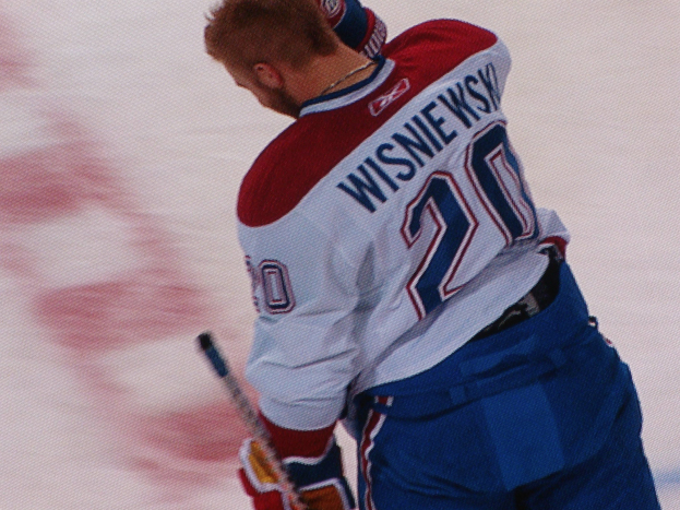 Ein Eishockeyspieler in einem "Wisniewski"-Trikot steht auf dem Eis und hölt einen Eishockeyschläger, mit einem konzentrierten und entschlossenen Gesichtsausdruck.