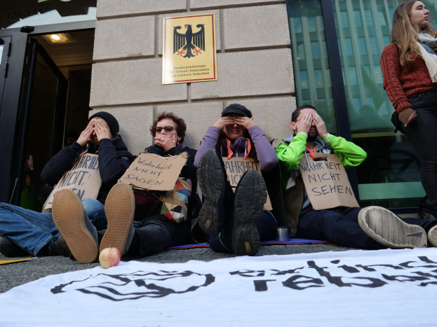 Eine Gruppe von Menschen sitzt vor einem Gebäude auf dem Boden und hält Schilder hoch, mit einer Fahne und einem Apfel auf der Straße und einer Frau, die rechts steht und besorgt aussieht.