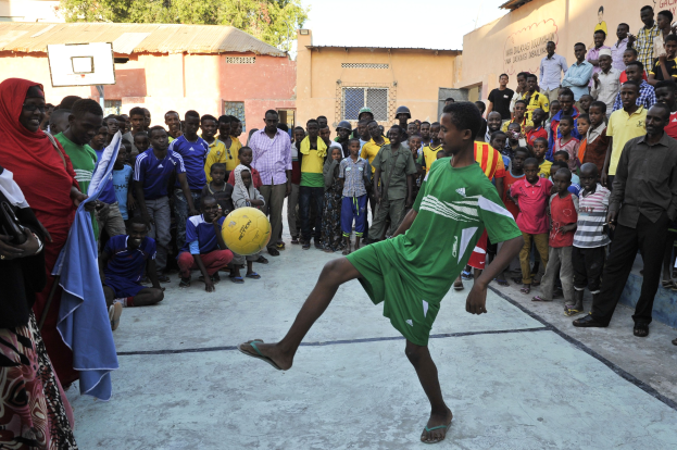 Ein junger Junge kickt einen Fuball vor einer Menge von Menschen, einige stehen und einige sitzen auf dem Boden, mit Gebuden, Bumen und einem klaren blauen Himmel im Hintergrund.