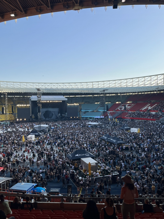 Große Menschenmenge sitzt und steht in einem Stadion mit Zelten, Geländern und Deckenlampen unter einem Dach, mit dem Himmel im Hintergrund.