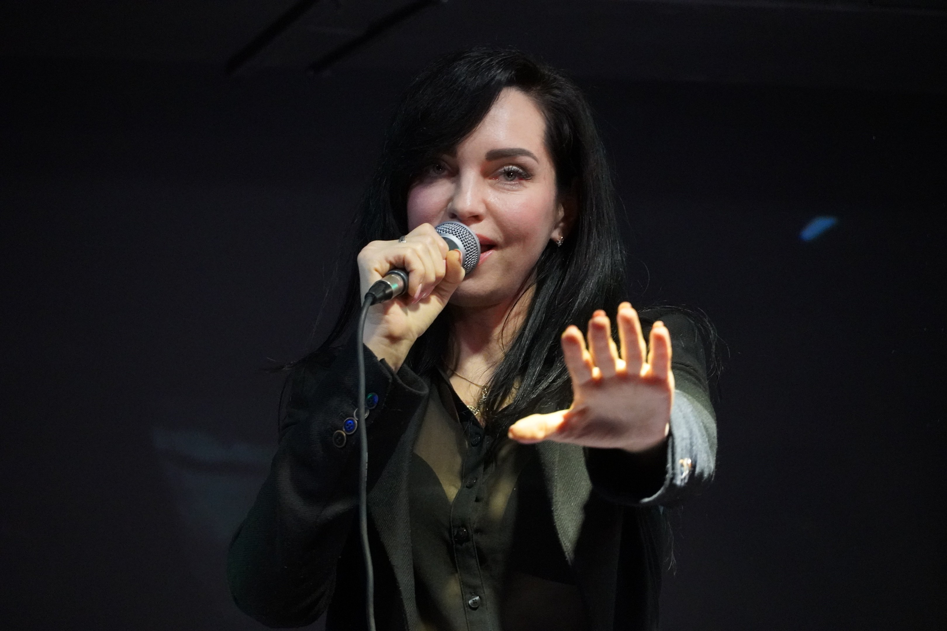 Eine Frau in einem schwarzen Kleid mit einem Dutt singt leidenschaftlich in ein Mikrofon auf einer hell erleuchteten Bühne, ihre Augen geschlossen und ihr Gesichtsausdruck intensiv.