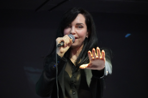 Eine Frau in einem schwarzen Kleid mit einem Dutt singt leidenschaftlich in ein Mikrofon auf einer hell erleuchteten Bühne, ihre Augen geschlossen und ihr Gesichtsausdruck intensiv.