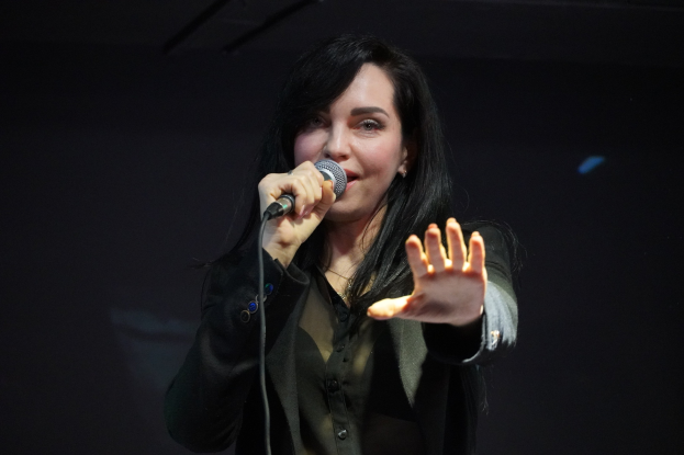 Eine Frau in einem schwarzen Kleid mit einem Dutt singt leidenschaftlich in ein Mikrofon auf einer hell erleuchteten Bühne, ihre Augen geschlossen und ihr Gesichtsausdruck intensiv.