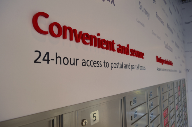 Wandschild wirbt für 24-Stunden-Zugang zu sicheren Post- und Paketfächern, mit nummerierten Fächern darunter sichtbar.