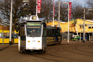 Eine weiße und blaue Straßenbahn auf einer Stadtstraße mit einer Person darin, umgeben von Laternenmasten, Bannern, einem Zaun, Fahrzeugen, Gebäuden, Bäumen und einem klaren blauen Himmel.