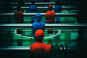 Ein buntfarbiger Kicker-Tisch mit einer Gruppe von Menschen in verschiedenenfarbigen Trikots, die in der Mitte des Tisches positioniert sind und bereit sind, ein Kicker-Spiel zu beginnen.