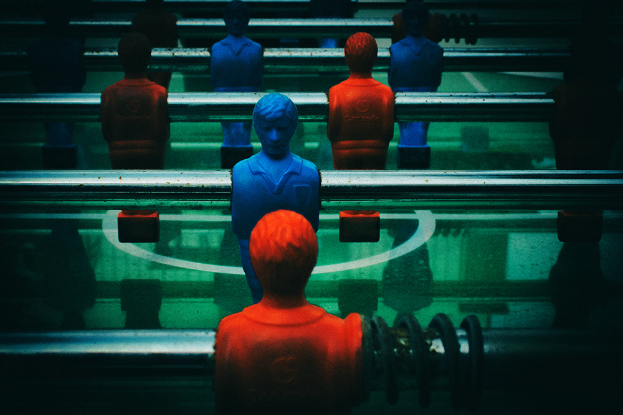 Ein buntfarbiger Kicker-Tisch mit einer Gruppe von Menschen in verschiedenenfarbigen Trikots, die in der Mitte des Tisches positioniert sind und bereit sind, ein Kicker-Spiel zu beginnen.