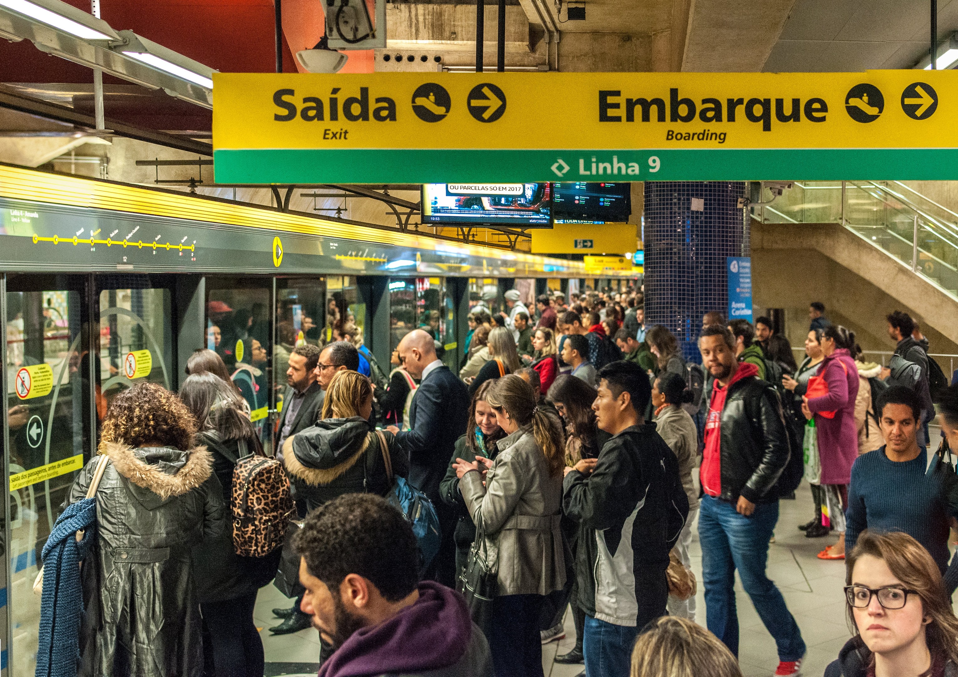 Eine Menschenmenge steht in einer U-Bahn-Station mit einem Zug auf der linken Seite, beleuchtet von Deckenlampen.