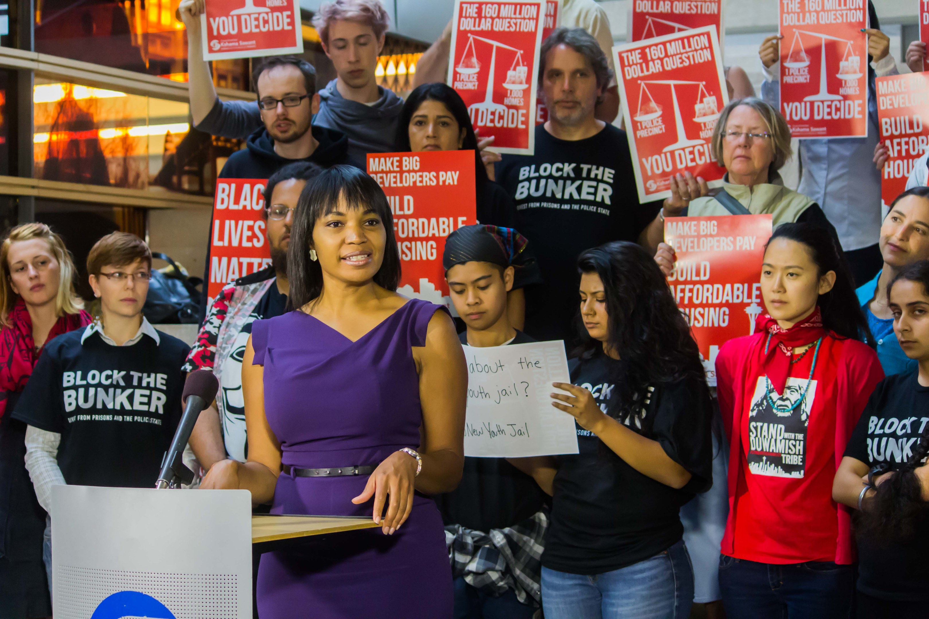 Eine Black-Lives-Matter-Aktivistin spricht in ein Mikrofon an einem Rednerpult zu einer Menge mit Schildern, mit Scheinwerfern und Equipment im Hintergrund.