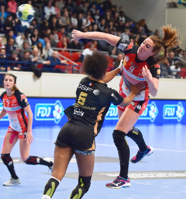 Eine Gruppe von Frauen spielt Handball auf einem Feld mit einem Ball in der Luft, in einer Stadionumgebung mit Zuschauern im Hintergrund.