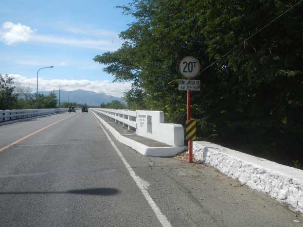 Fahrbahn mit Fahrzeugen, Geschwindigkeitsbegrenzungsschild, Geländer, Laternen, Bäume und bewölkter Himmel im Hintergrund.