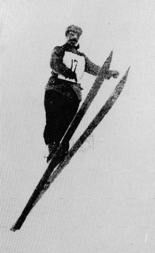 Schwarzes und weißes Foto eines Skifahrers in der Luft, der Skier hält, eine Skiausrüstung trägt und einen entschlossenen Gesichtsausdruck hat.