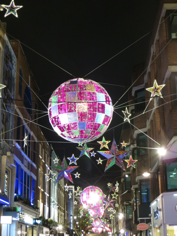 Eine nächtliche Stadtstraße mit zahlreichen Lichtern und Dekorationen, darunter eine große Diskokugel, die von einem Gebäude hängt und eine festliche Atmosphäre schafft.