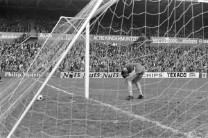 Schwarzes und weißes Foto eines Fußball-Torwarts, der einen Ball in der Nähe eines Torpfostens hält, mit Zuschauern im Hintergrund.
