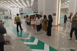 Menschen in einer Reihe an einem Flughafen-Sicherheits-Check-in, mit Taschen und Mobiltelefonen, mit Säulen und Deckenleuchten im Hintergrund.