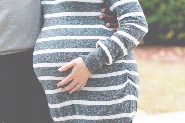 Schwangeres Paar in Umstandskleidung, das zusammen steht und den Bauch hält, umgeben von Bäumen im Hintergrund.