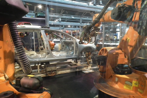 Ein Auto in einer Fabrik mit einem Roboterarm auf der rechten Seite, andere Autos auf der Fertigungsstraße im Hintergrund und eine beleuchtete Decke.