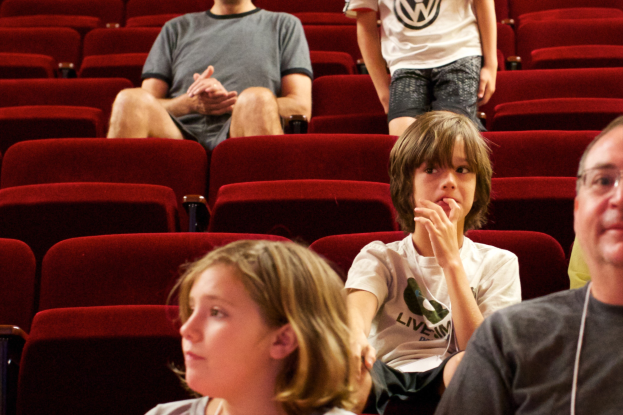 Eine Gruppe von Menschen in einem Kinosaal mit roten Stühlen, ein Junge in einem weißen T-Shirt und schwarzen Hosen in der Mitte, umgeben von anderen in verschiedenenfarbiger Kleidung.