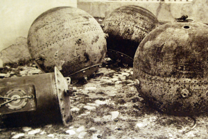 Drei große Metallbomben liegen auf dem Boden vor einer Wand mit einem Fenster im Hintergrund, dargestellt in einem Schwarz-Weiß-Foto.