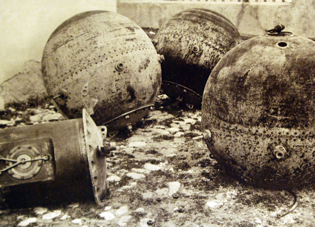 Drei große Metallbomben liegen auf dem Boden vor einer Wand mit einem Fenster im Hintergrund, dargestellt in einem Schwarz-Weiß-Foto.