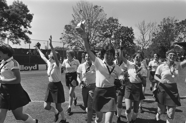Eine Gruppe von Frauen läuft auf einem Rasenfußballfeld, jede hält einen Pokal, mit Bäumen, Maschendrahtzaun und Texttafeln im Hintergrund, dargestellt in Schwarz-Weiß.