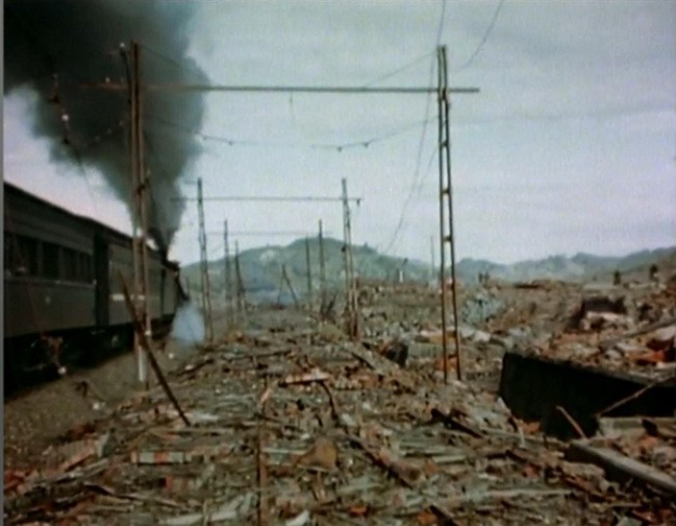 Ein Zug fährt durch ein zerstörtes Gebiet mit Rauch, der aus ihm aufsteigt, umgeben von Strommasten und Drähten, Hügeln und einem klaren Himmel im Hintergrund und verstreutem Schutt auf dem Boden.