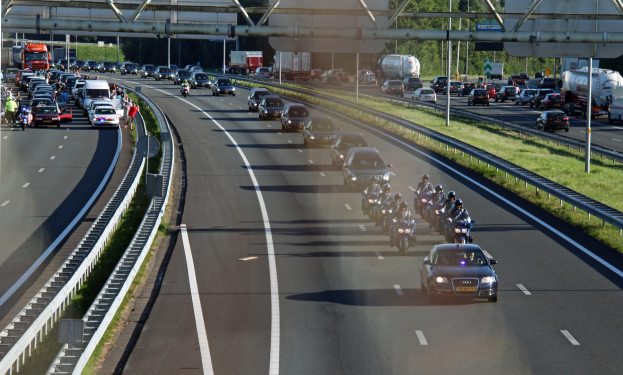 Eine vielbefahrene Autobahn mit zahlreichen Autos und Motorrädern, Geländer auf beiden Seiten, Gras am Boden und ein Hintergrund mit Bäumen, Pfählen, Texttafeln und einer Brücke mit Stangen.