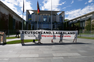 Eine Gruppe von Menschen hält ein Transparent mit der Aufschrift "Deutschland, Lagerland Migration ist kein Verbrechen" vor einem eingezäunten Gebäude mit Bäumen und Fahnen.