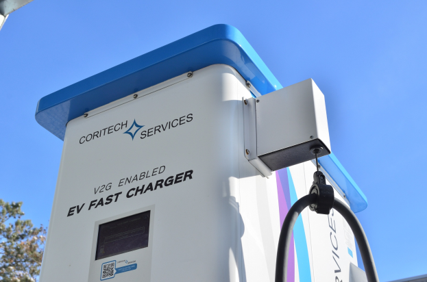 Elektrofahrzeug-Ladestation mit der Beschriftung "V2G Fast Charger" mit B├Ąumen und einem Geb├Ąude im Hintergrund bei einem klaren blauen Himmel.