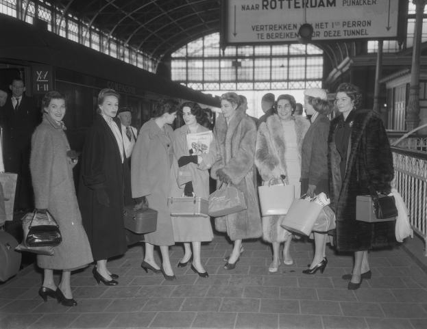 Schwarz-weißes Bild einer Gruppe von Frauen, die an einem Bahnhof stehen und Taschen halten, mit einem Zug auf der linken Seite und Geländern und einer Tafel auf der rechten Seite.