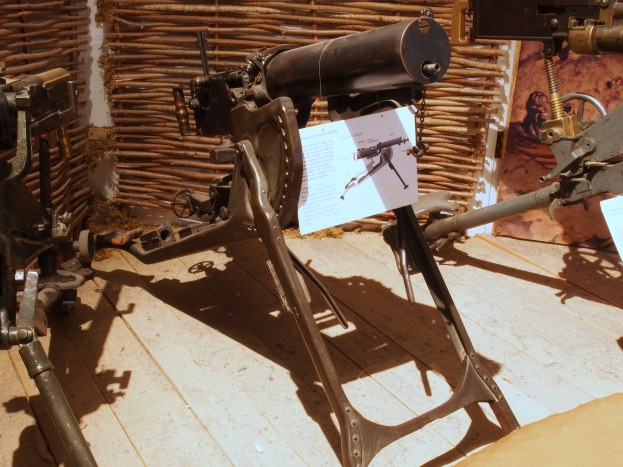 Eine Gatling Gun, eine Art Maschinengewehr, das von der britischen Armee während des Zweiten Weltkriegs verwendet wurde, wird auf einem hölzernen Boden in einem Museum ausgestellt, mit einer Wand im Hintergrund und einer Texttafel rechts.