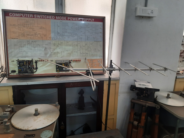 Ein unordentlicher Raum mit verschiedenen Elektronikgeräten, darunter eine beschriftete Platine auf einem Tisch, Metallstangen, eine wandmontierte Verteilerbox, ein Fenster und Gegenstände in Schränken, die auf eine computergeschaltete Modus-Stromversorgung hinweisen.