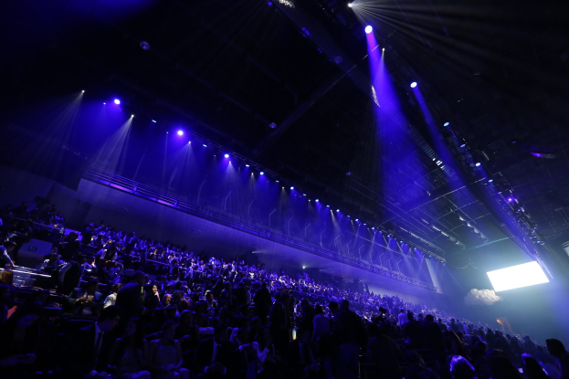 Gro├če Menge in einem abgedunkelten Konzertsaal vor einer Bühne mit Deckenlichtern und einem Bildschirm auf der rechten Seite.