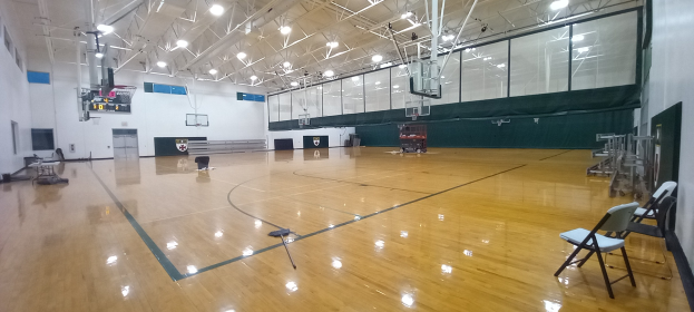 Großer Turnsaal mit Holzboden, Stühlen, einer Wand, Deckenleuchten und einem Basketballfeld.