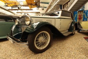 Ein Oldtimer-Auto in einem Museum zwischen anderen Fahrzeugen ausgestellt, mit einem Textschild rechts daneben und Flugzeugen an der Decke im Hintergrund.