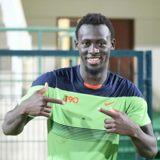 Ein Mann in einem grün-blauen Hemd, der lachend in die Kamera zeigt und im Hintergrund eine undeutliche Brüstung, Wand und Tür zu sehen ist.