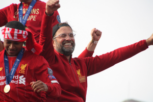 Liverpool-Trainer Jürgen Klopp feiert mit Spielern und Staff nach dem Gewinn der UEFA Champions League, trägt eine rote Jacke mit zwei Medaillienträgern und einem sichtbaren Himmel im Hintergrund.