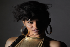 Eine Frau in einem goldenen Kleid und großen Ohrringen steht selbstbewusst vor einer Wand, ihr glatter Bob und das natürliche Make-up unterstreichen ihren entschlossenen Gesichtsausdruck, während sie direkt in die Kamera schaut.