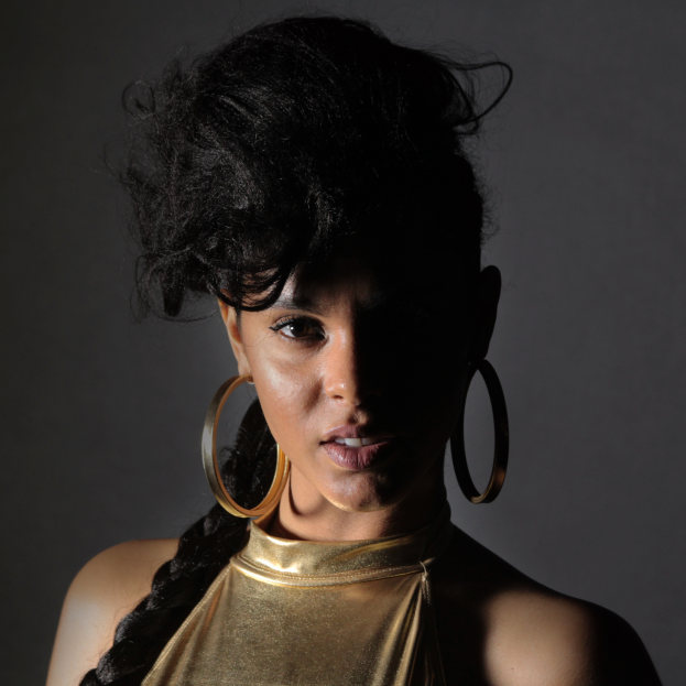 Eine Frau in einem goldenen Kleid und großen Ohrringen steht selbstbewusst vor einer Wand, ihr glatter Bob und das natürliche Make-up unterstreichen ihren entschlossenen Gesichtsausdruck, während sie direkt in die Kamera schaut.