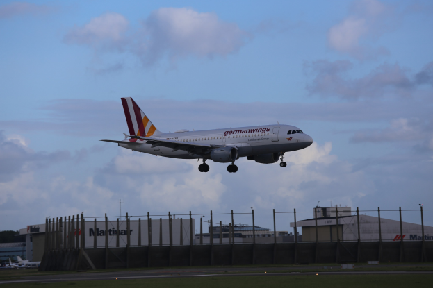 Germanwings Airbus A320-200 beim Abheben vom Frankfurt Airport mit Gras darunter, einem Zaun im Vordergrund, Gebäuden und Bäumen im Hintergrund und einer bewölkten Himmel darüber.