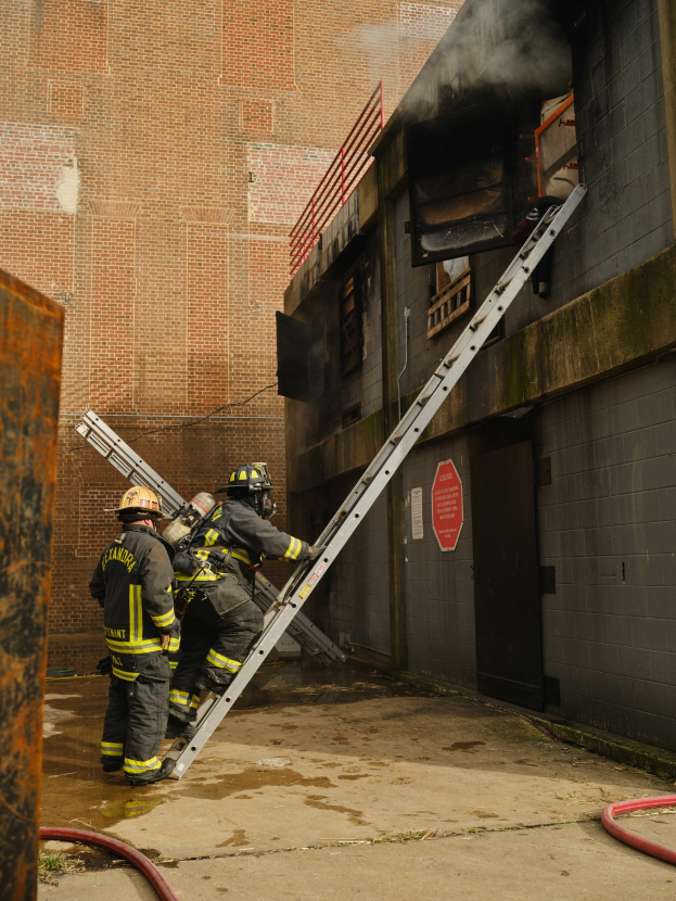 Feuerwehrleute in Helmen, die eine Leiter zu einem raucherfüllten Gebäude hinaufsteigen.