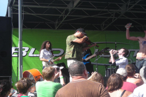 Eine Gruppe von Musikern performs auf der Bühne mit einem Publikum, das eine große schwarze Box auf der linken Seite und ein grünes Banner im Hintergrund zeigt.