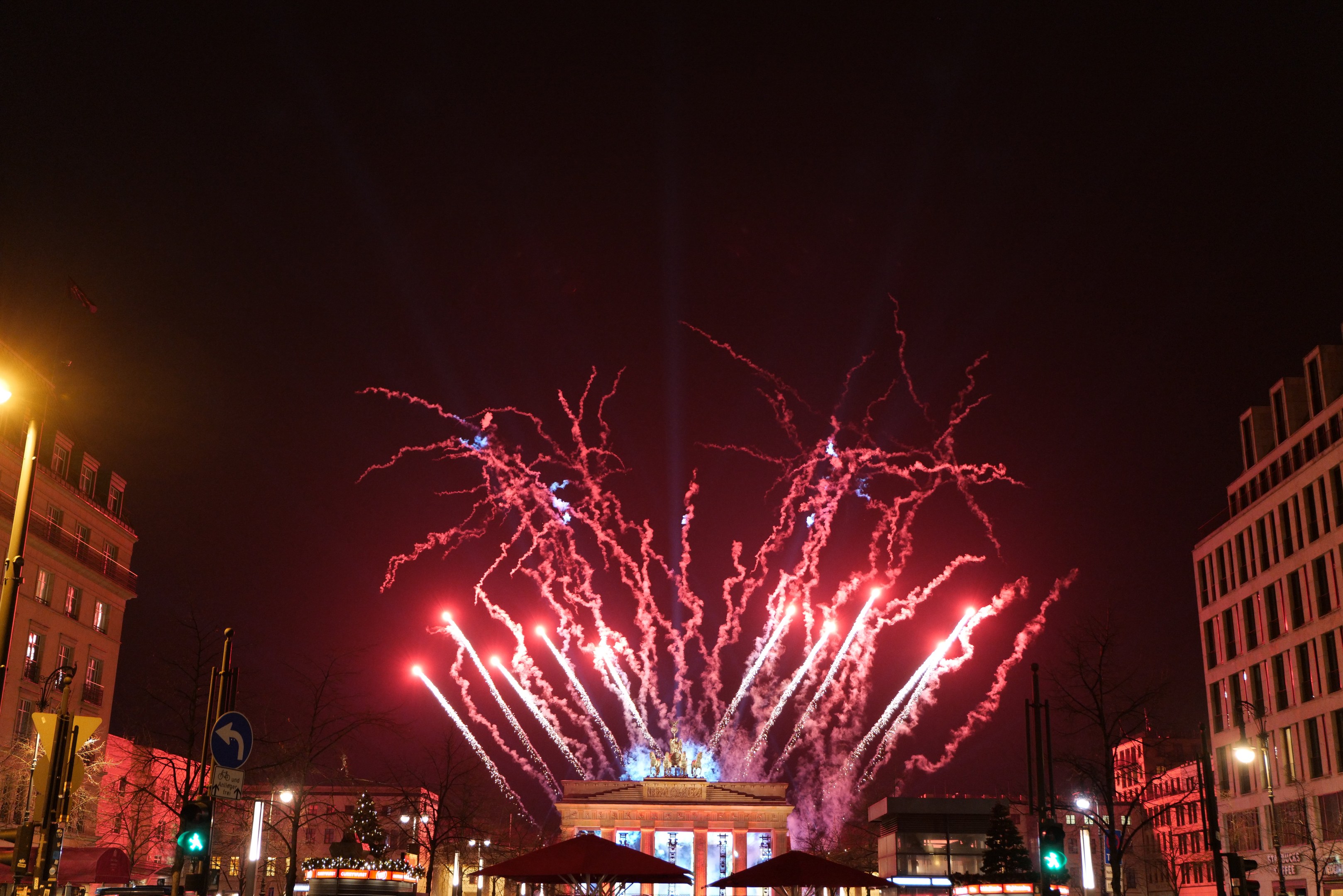 Eine belebte Stadtstraße an Silvester in Berlin, voller Menschen, Fahrzeuge und Gebäude, beleuchtet von Lichtern und Feuerwerk am Himmel.