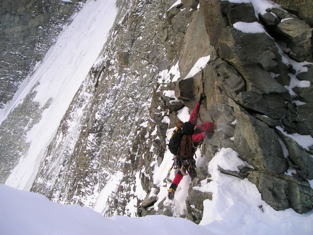 Eine Person in einer roten Jacke klettert einen steilen, schneebedeckten Berghang hinauf und trägt eine Tasche.